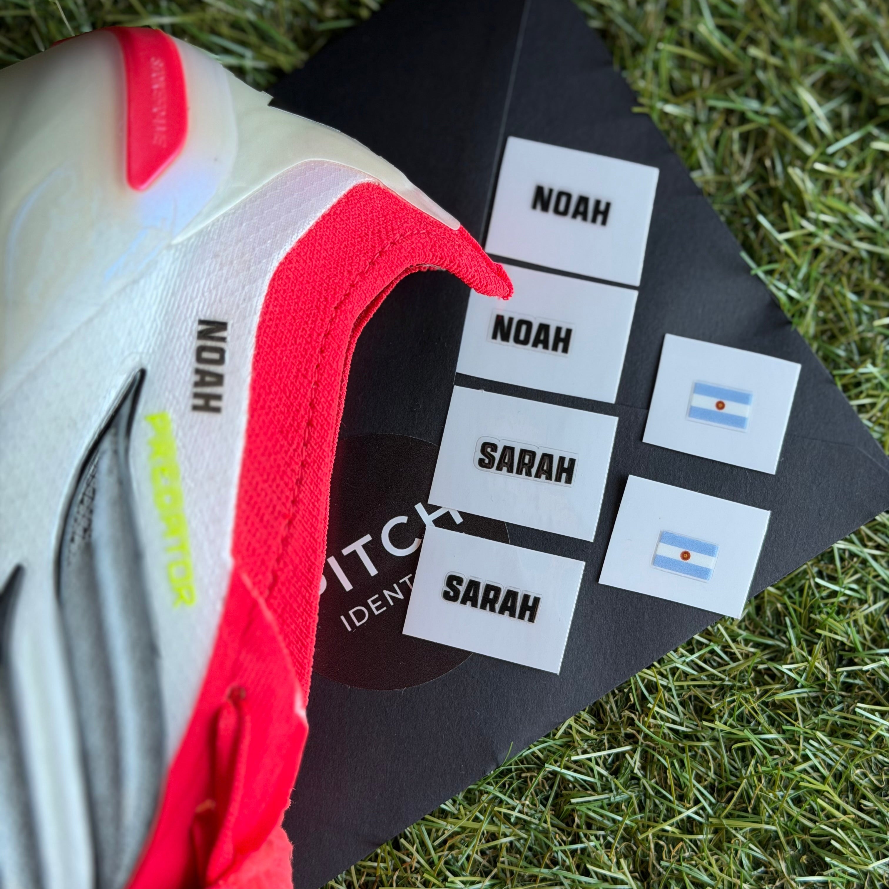 A close-up of a white and red athletic shoe resting on a black printed card placed on green grass. The card shows the names NOAH and SARAH in black text on white labels, alongside two small Argentina flag stickers.