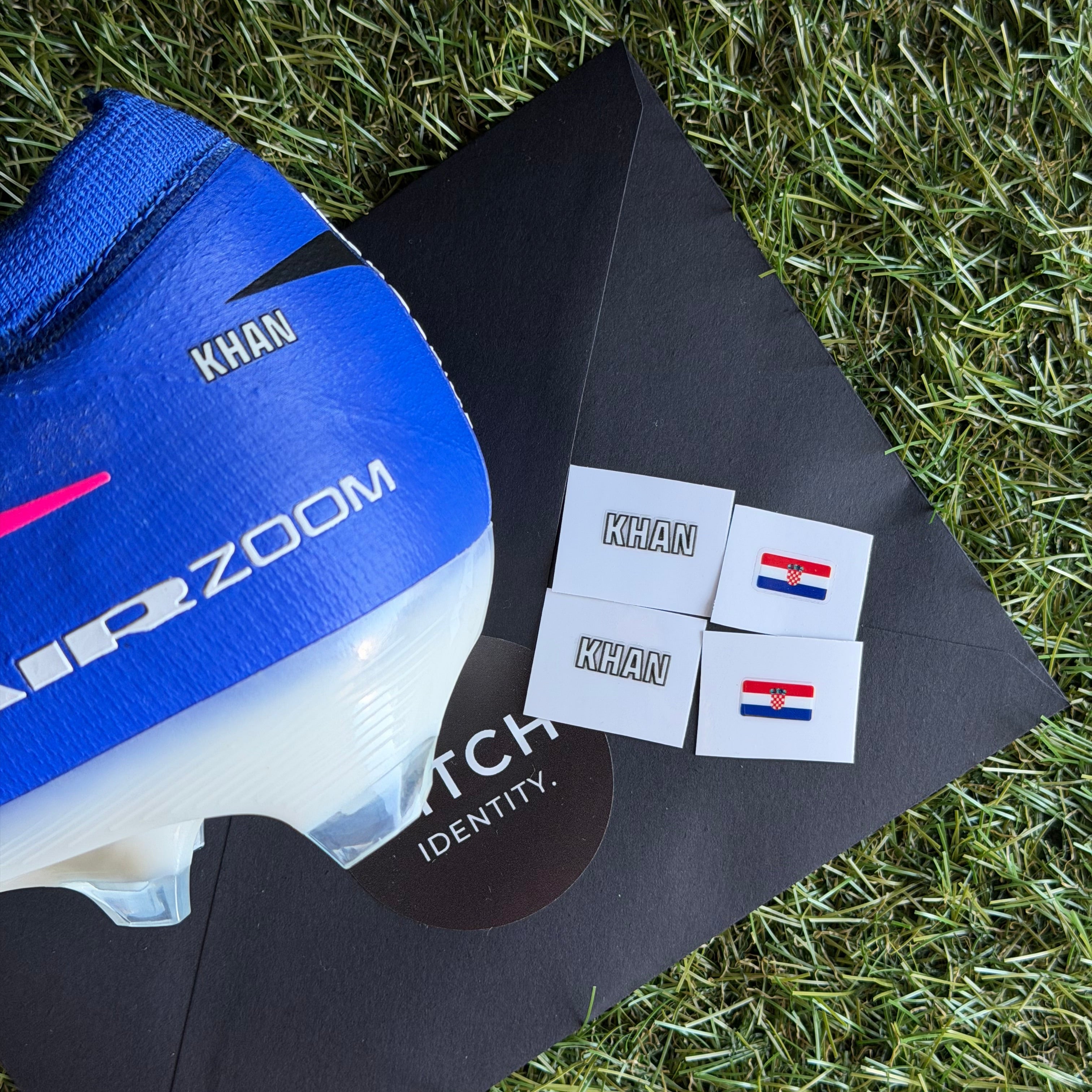 A blue and white Nike Air Zoom soccer cleat is displayed on a black Nike box on green grass, next to four small white patches featuring the name 'KHAN' and the Netherlands flag.